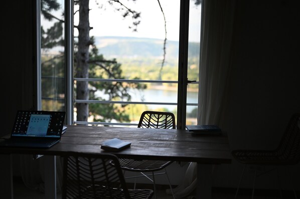 A room with a wooden table, a laptop, and a chair. A window offers a view of trees and a body of water.