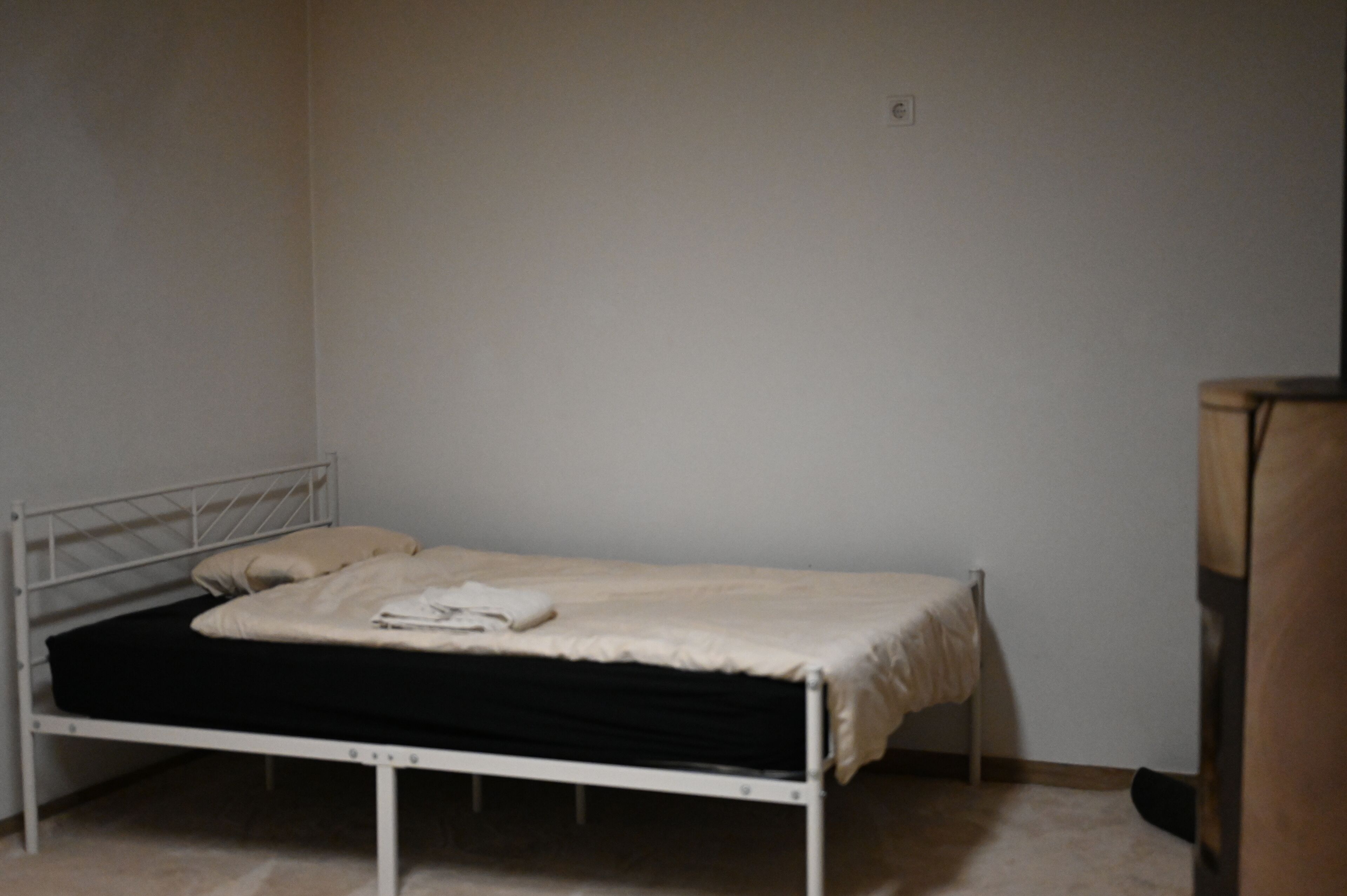 A single bed with a white mattress and a black bottom, a white blanket, and a pillow on a metal frame bed in a room with plain walls and a wooden piece of furniture to the right.