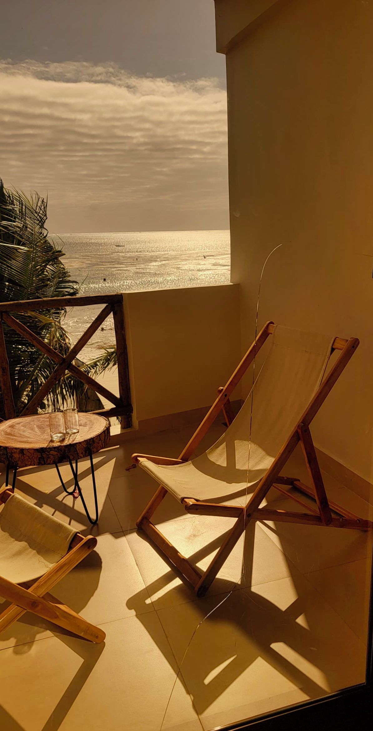 A balcony with a view of the sea, two wooden lounge chairs, and a small round table with glasses.