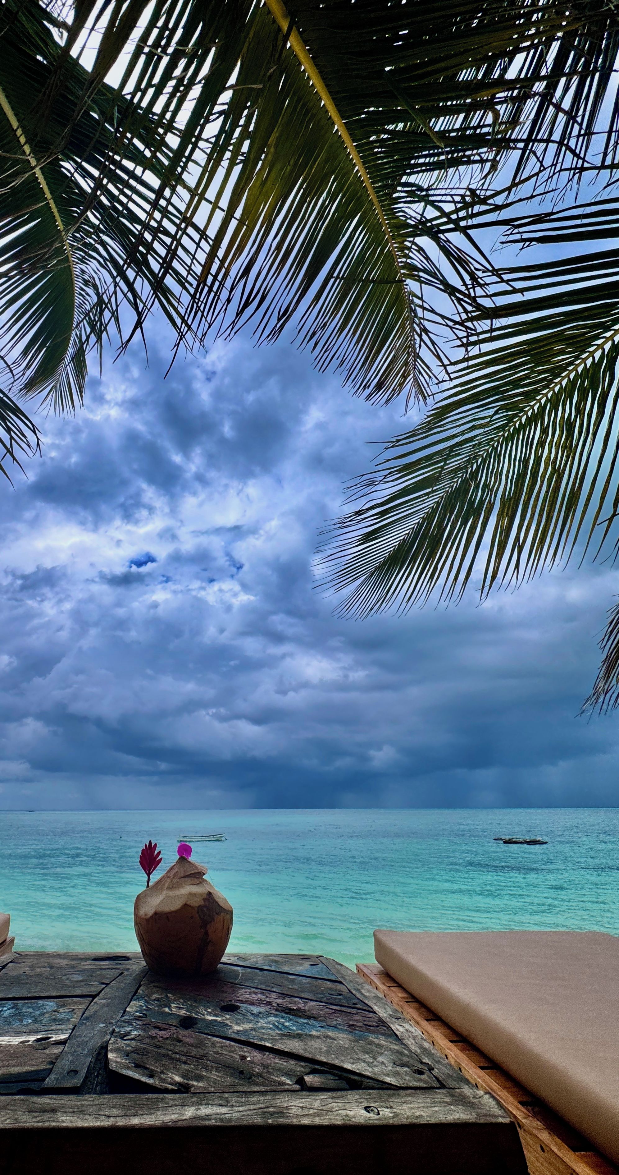 A wooden table with a coconut and a decorative item, overlooking a turquoise sea under a cloudy sky.