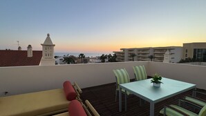 Una terraza con mesa y sillas, un sofá, y vista al mar y edificios al atardecer.