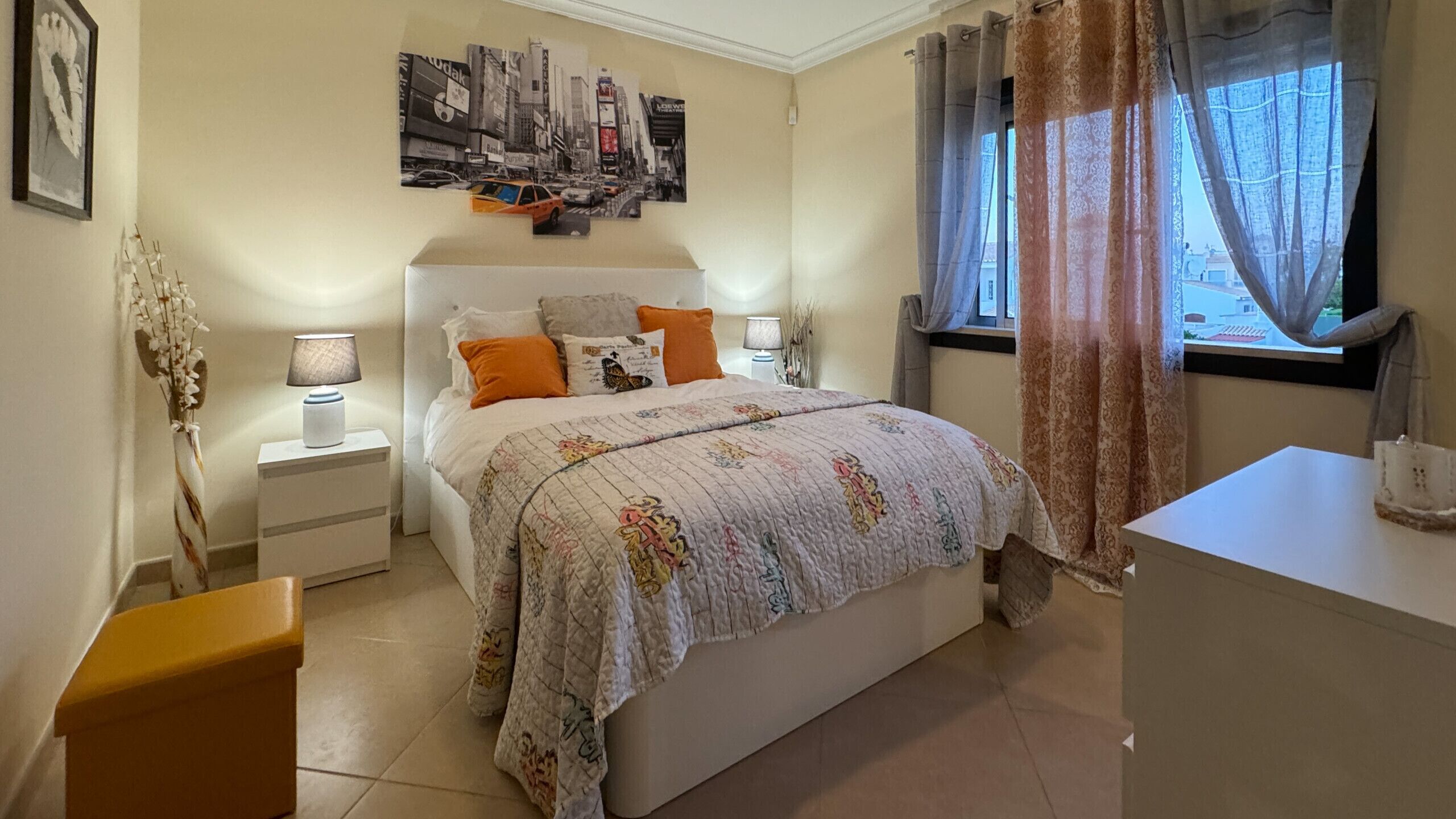 Un dormitorio con cama, mesitas de noche, cómoda, ventana con cortinas y un cartel con un paisaje urbano en la pared.