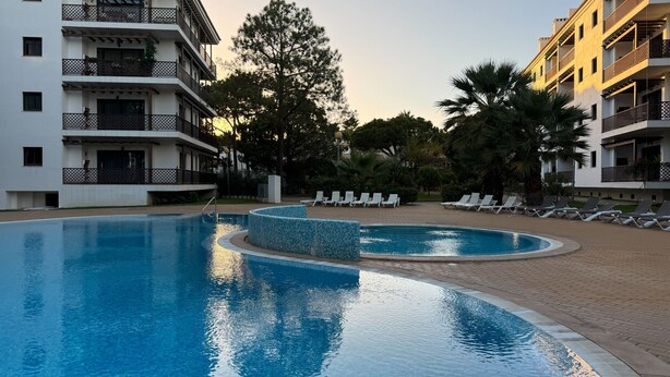 Una piscina con borde de baldosas, rodeada de sillones de descanso y un edificio con balcones.