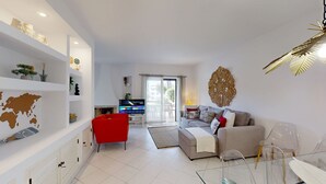 Un salon moderne avec un fauteuil rouge, un canapé gris et un téléviseur posé sur un meuble. On y trouve des étagères ornées d’objets décoratifs et un grand élément décoratif mural.