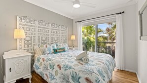 Un dormitorio con una cama, dos mesitas de noche, un ventilador de techo y vistas a un balcón con plantas.