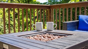A wooden deck with two white mugs on a table, a fire pit, and a wicker chair with a blue cushion.