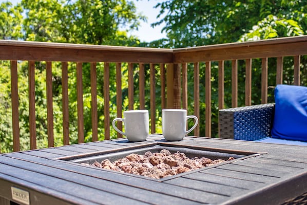 Une terrasse en bois avec deux tasses blanches posées sur une table, un foyer et une chaise en osier dotée d’un coussin bleu.