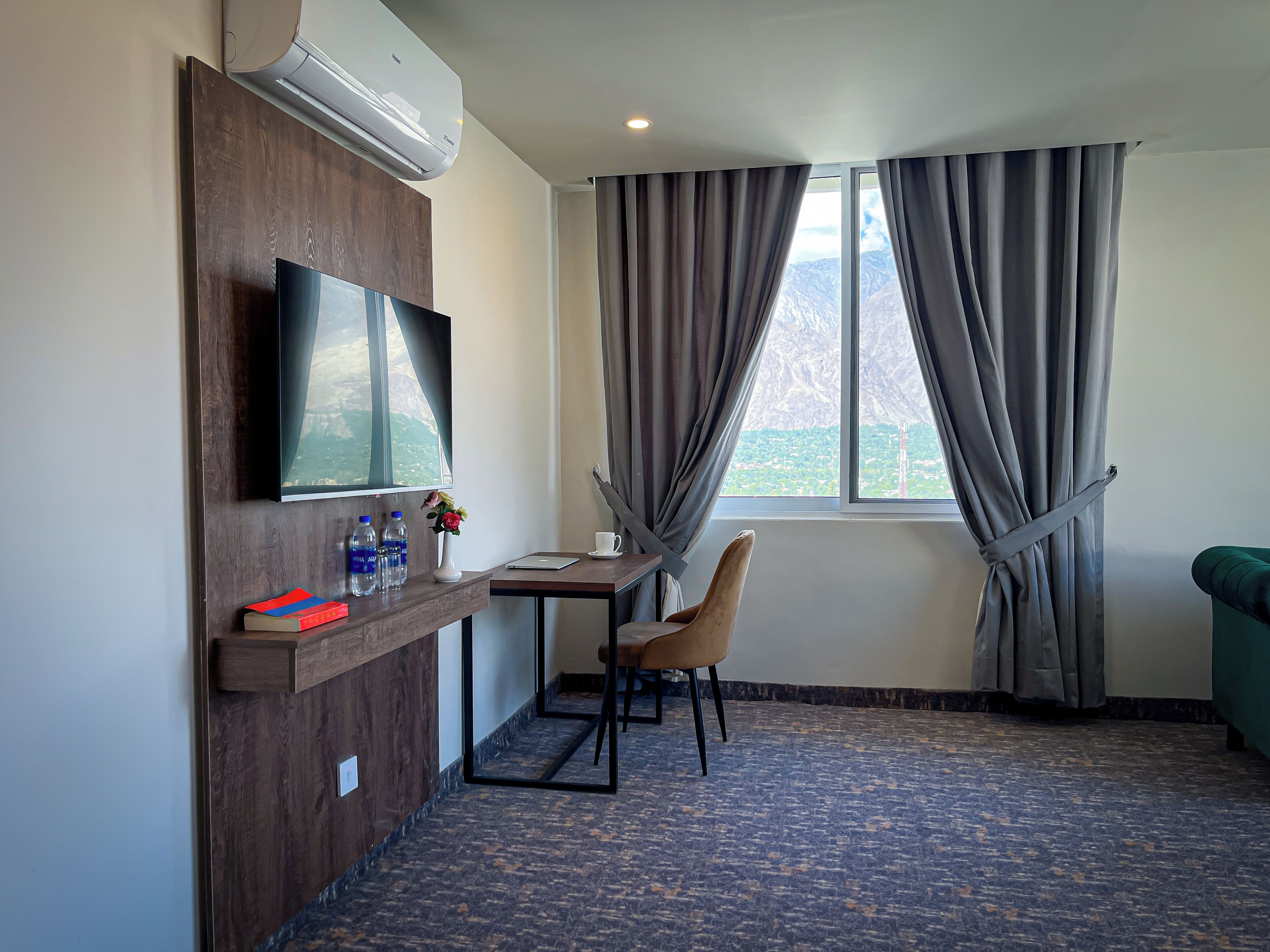 A hotel room with a flat-screen TV mounted on the wall, a wooden desk with a laptop, a chair, a small table with a cup, and a view of mountains through the window.