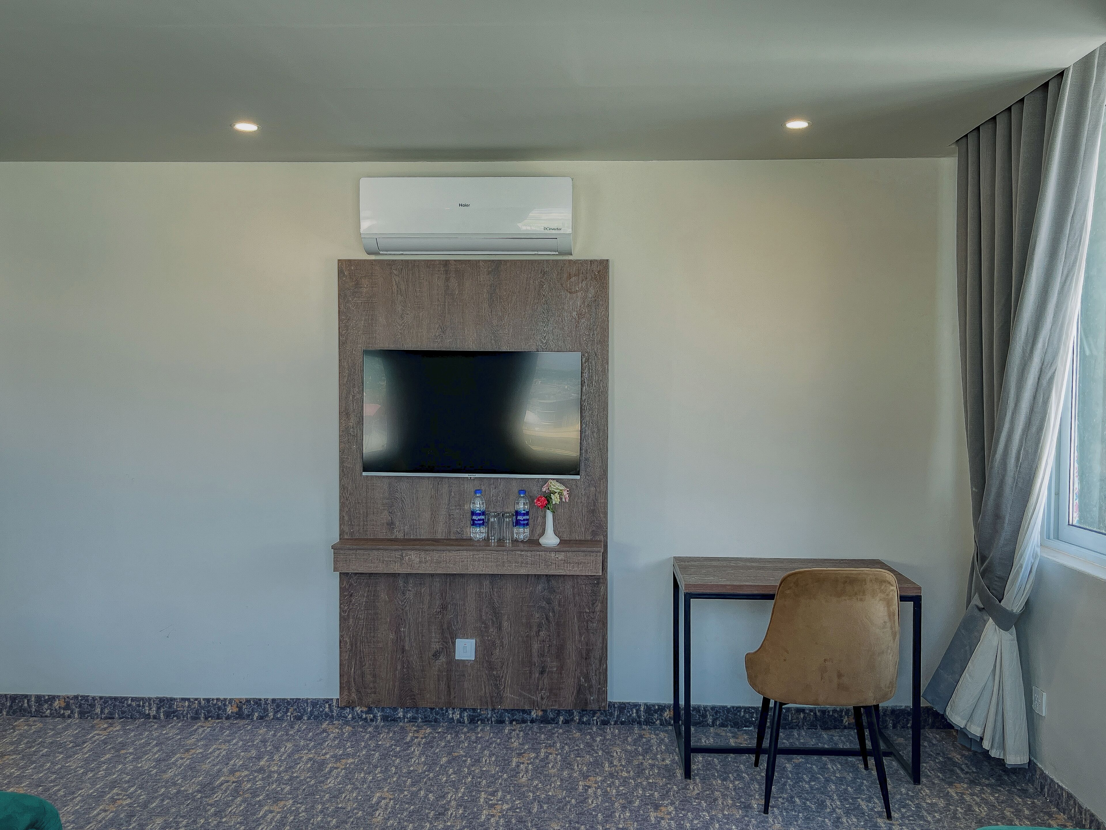 A hotel room with a flat-screen TV mounted on a wooden panel, a small table with a chair, and a window with curtains.