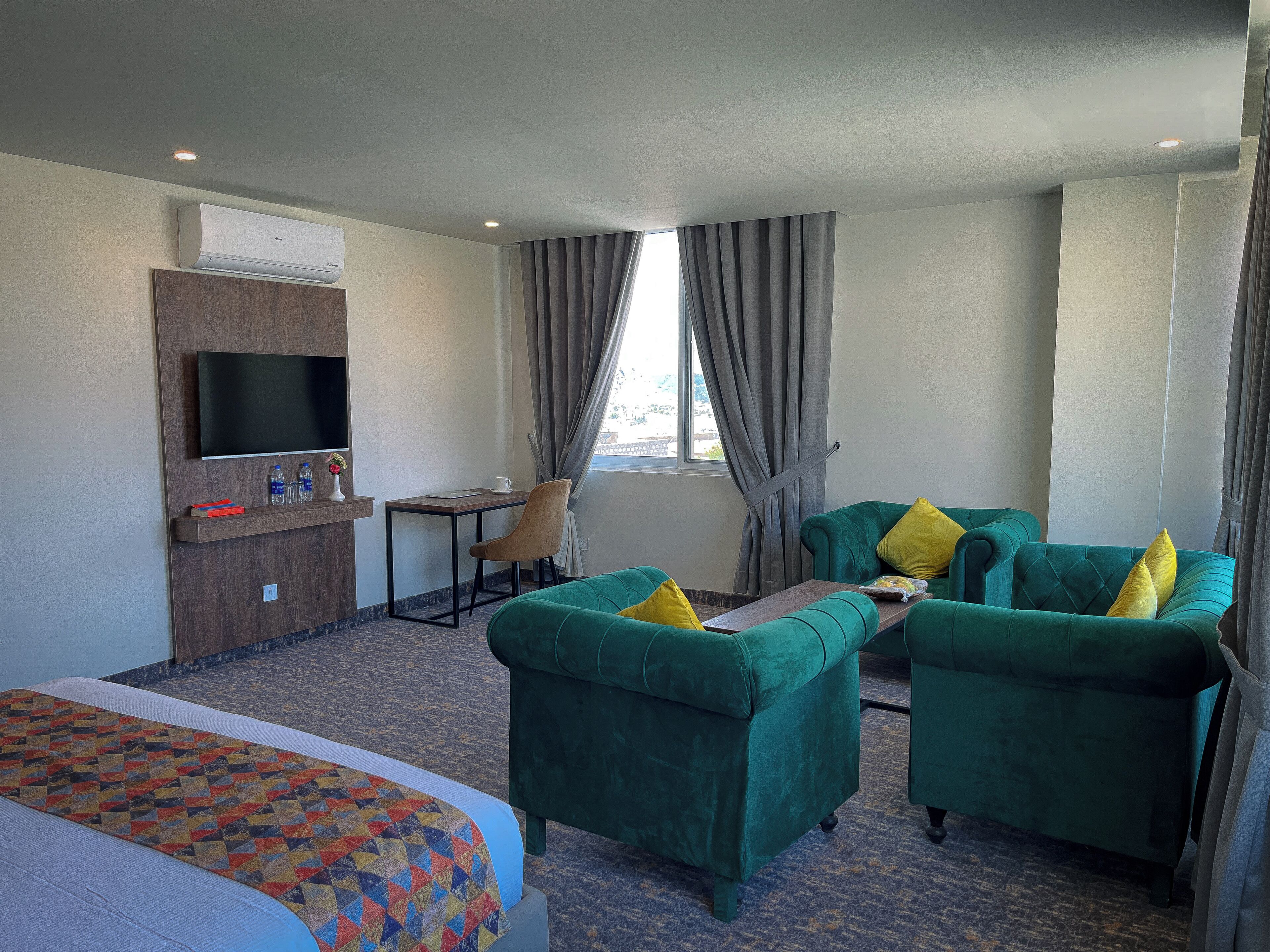 Habitación de hotel con cama, escritorio, silla, dos sofás verdes con almohadas amarillas, televisor de pantalla plana y ventana con cortinas.