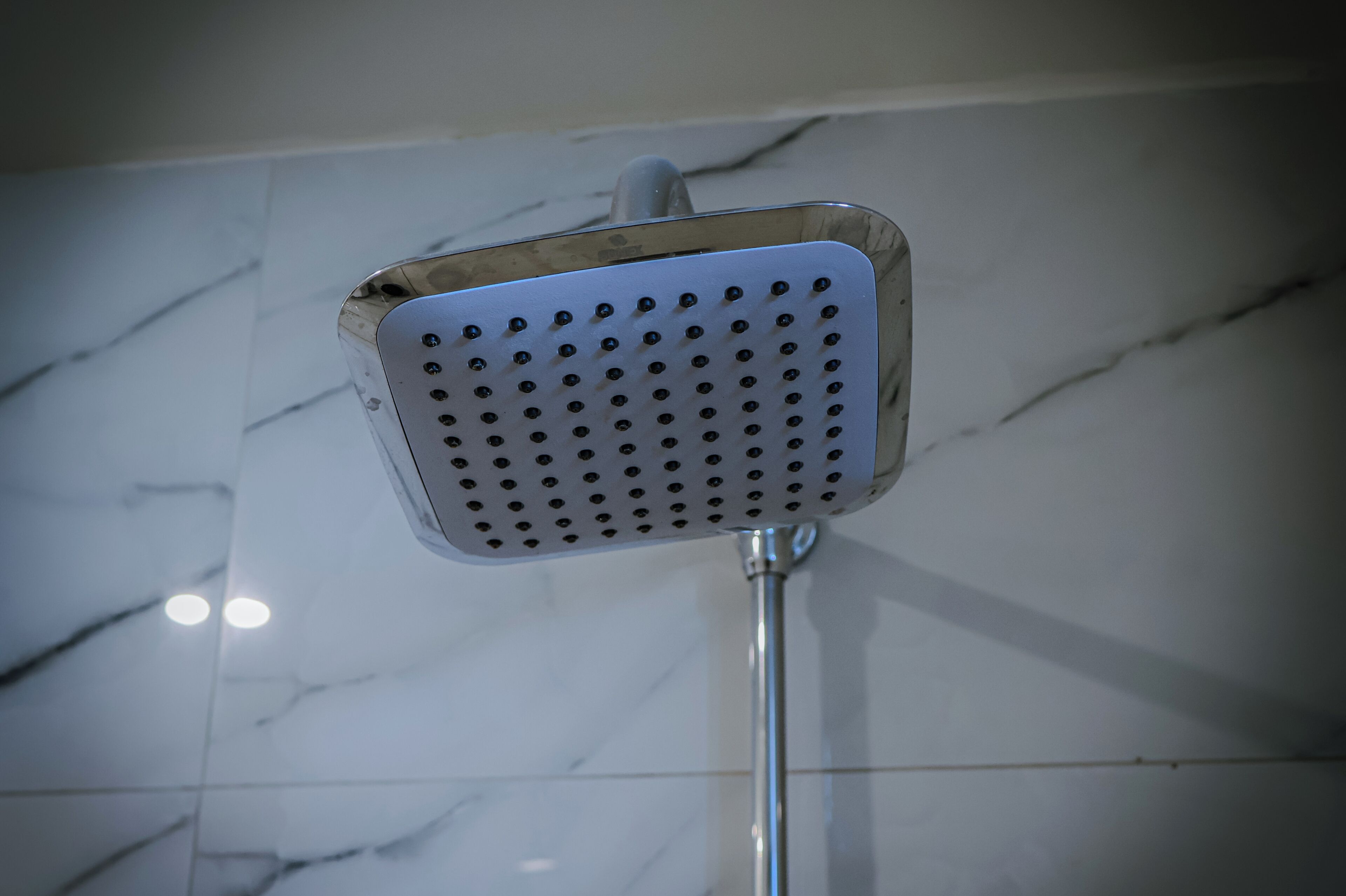 A showerhead with a square design and multiple holes, mounted on a metal pole, against a tiled wall.