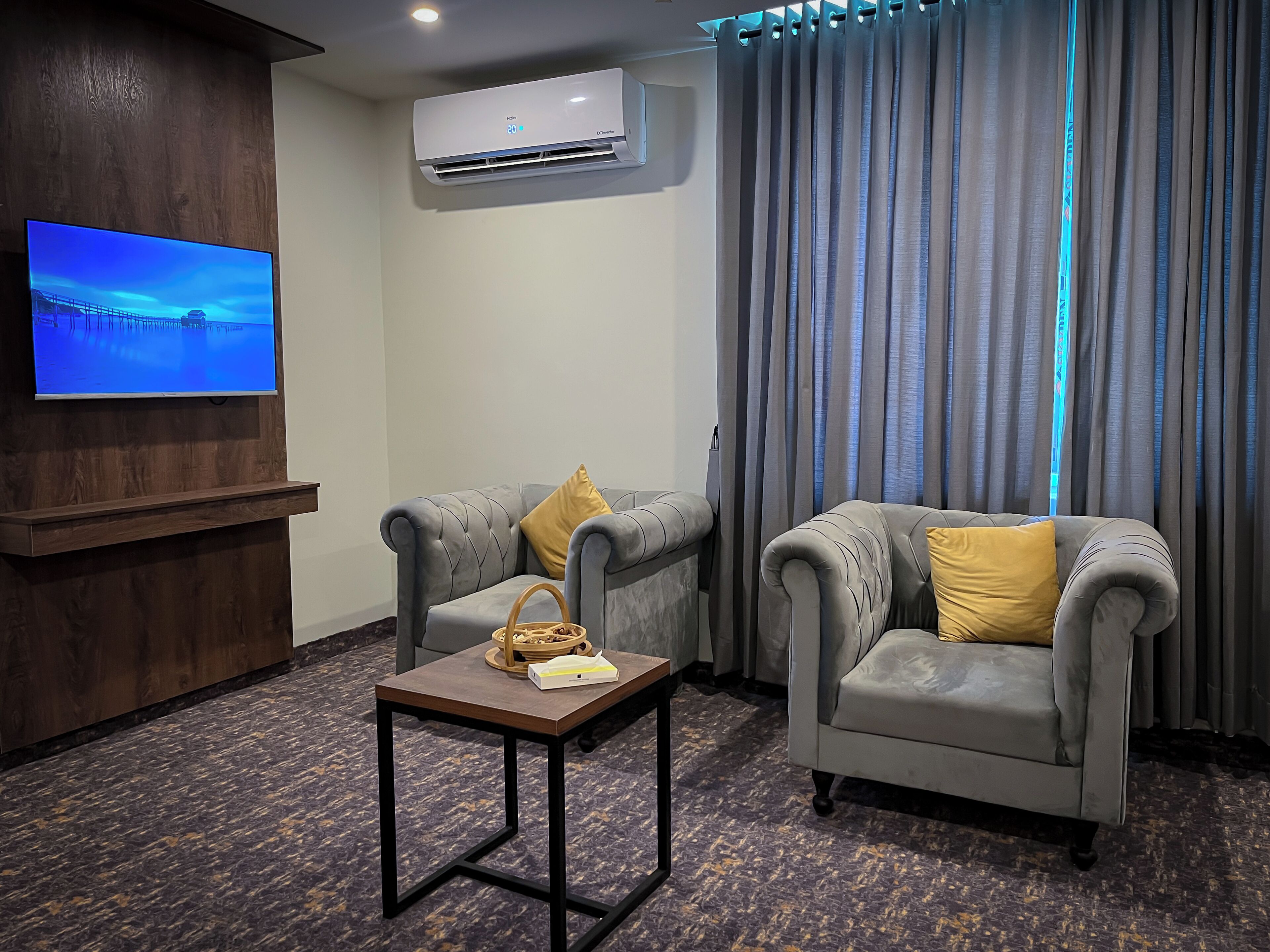 A hotel room with a flat-screen TV, two grey armchairs with yellow cushions, a small table with a basket and a box, and a large window with grey curtains.