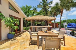 An outdoor patio with a wooden table and chairs, a thatched-roof bar area, and potted plants.