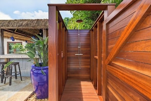 A wooden outdoor shower area with a thatched roof, a potted plant, and a wooden fence.