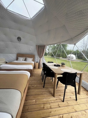 A cozy interior with a wooden floor, a bed, a table with chairs, and a view of the outdoors through a geometric window. - Grand Luxe Geodome Glamping w/ 2 Queen beds North (Miller Lake)
