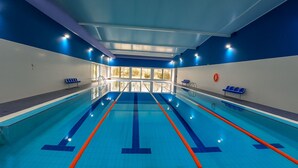 An indoor swimming pool with clear lanes, windows, and blue seating areas.