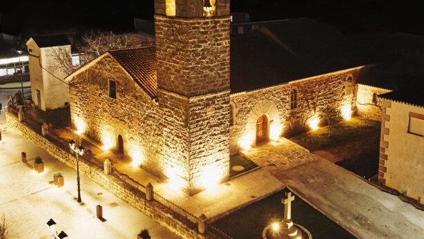 A stone church with a bell tower, illuminated by warm lights, and a fountain in the foreground.