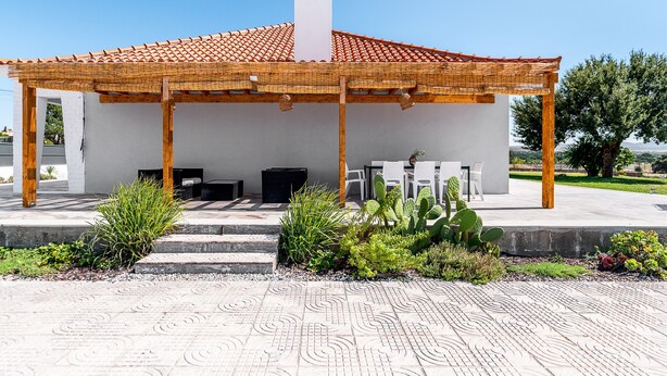 Een overdekt zitgedeelte buiten, met een pergola, een witte muur en een cactustuin.