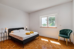 A bedroom with a bed, a bedside table with a lamp, a chair, and a window.