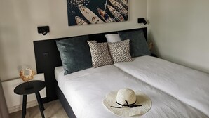 Una cama bien hecha con una colcha blanca y un sombrero de paja encima. En la pared sobre la cama, hay un cuadro de barcos.
