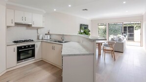 A modern kitchen with white cabinetry, a built-in oven, and a gas stove. There is a dining area with a table and chairs, and a large window providing a view of the outdoors. - Adelaide Oval Townhouse / Walk to the AFL & CBD (North Adelaide)