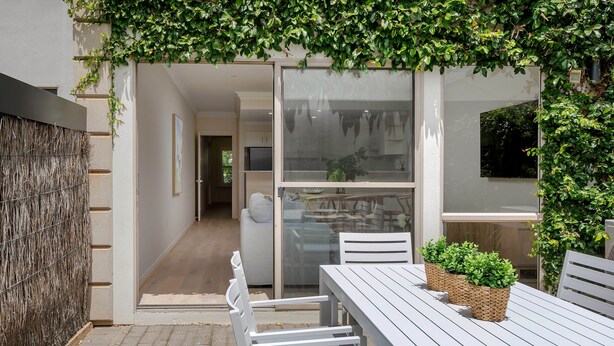 A modern outdoor patio with a white table and chairs, a woven wall, and a glass door leading to an interior space. - Adelaide Oval Townhouse / Walk to the AFL & CBD (North Adelaide)