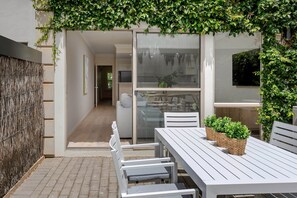 Patio esterno moderno con tavolo e sedie bianche, parete intrecciata e porta vetrata che dà su un ambiente interno.