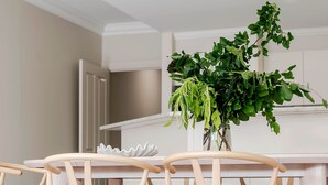 A dining area with wooden chairs and a table, a vase with green plants, and a white wall in the background. - Adelaide Oval Townhouse / Walk to the AFL & CBD (North Adelaide)