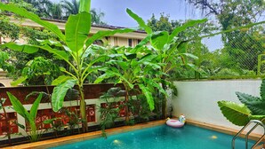 A swimming pool with a pink inflatable unicorn float, surrounded by lush greenery and a white wall. - ELIVAAS Mirai Tauras 3BHK (Vagator)