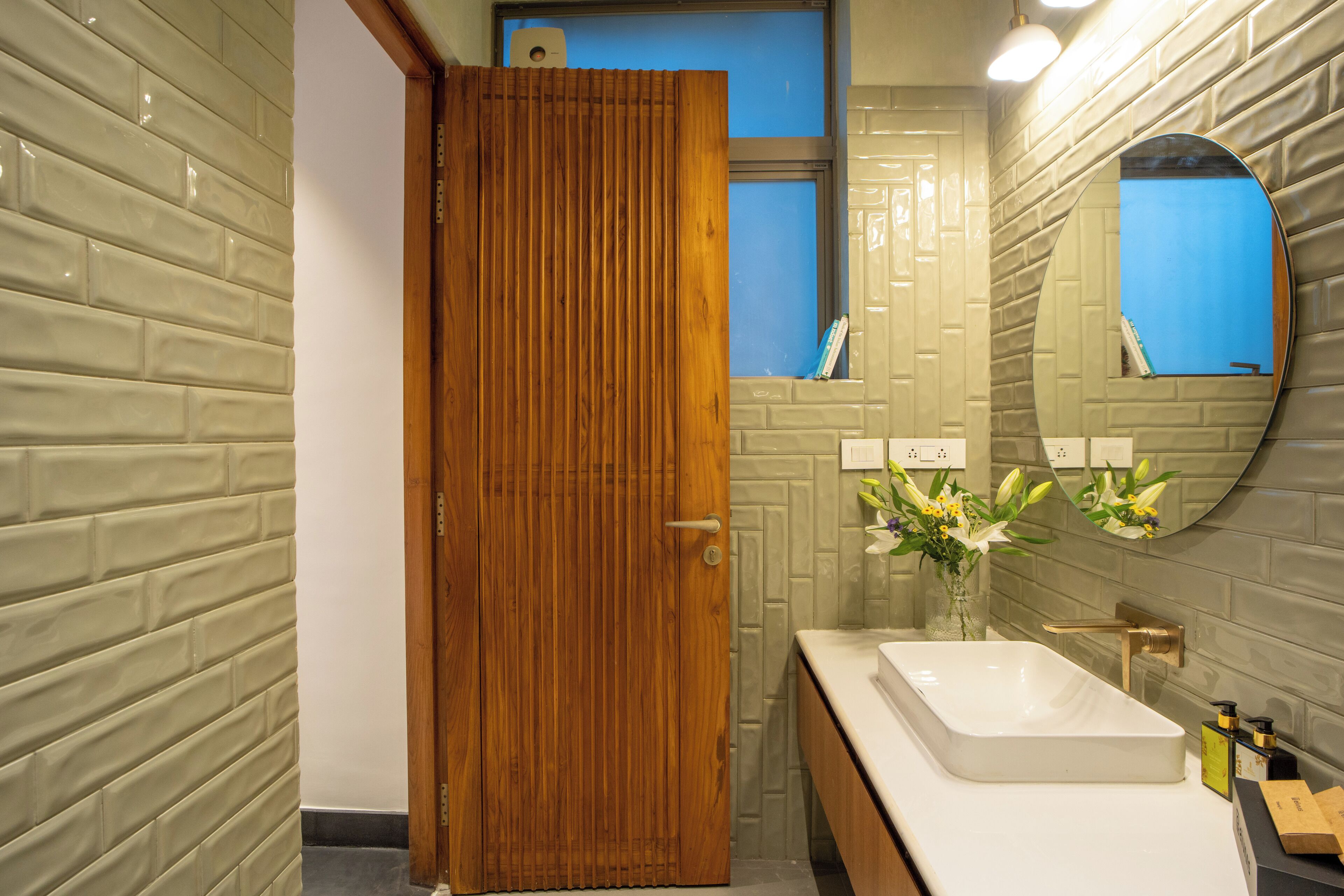 A bathroom with a wooden door, a white sink, a mirror, and a vase with flowers.
