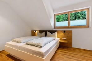 Een slaapkamer met een schuine kap, een bed met witte beddengoed, een houten hoofdeinde, een nachtkastje en een raam met uitzicht op bomen.