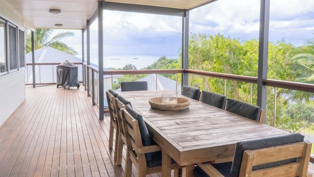 A covered outdoor dining area with a wooden table and chairs, overlooking a scenic view of the sea and greenery.