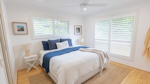 A bedroom with a bed, two bedside tables, a ceiling fan, and large windows with blinds.
