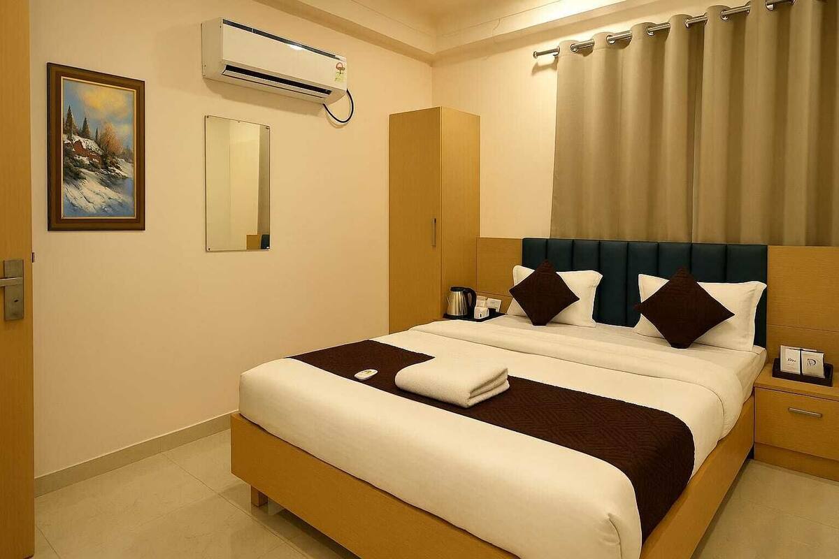 A hotel room with a bed, a bedside table, a nightstand, a mirror, an air conditioning unit, and a painting on the wall.