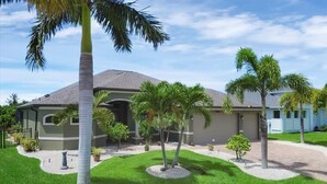 Una casa residencial con techo gris, rodeada por un césped cuidado y palmeras.