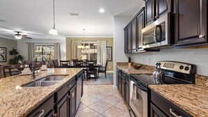 A modern kitchen with granite countertops, dark cabinetry, stainless steel appliances, and a dining area with a chandelier. - Cup8931 (Kissimmee)