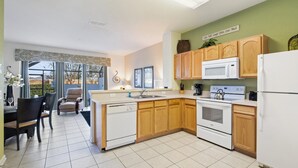 A modern kitchen with wooden cabinets, a white refrigerator, a microwave, and a stove. There is a dining area with a table and chairs, and a window with a view of greenery. - Msw2563 (Kissimmee)