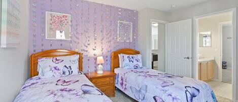 Un dormitorio con dos camas, cada una con una colcha de color púrpura con motivos de mariposas, una mesita de noche con lámpara y una pared decorada con pegatinas de mariposas.