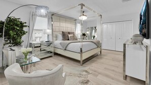 Een moderne slaapkamer met een hemelbed, een glazen salontafel, een witte fauteuil, een grote spiegel en een potplant.