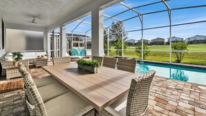 Een overdekt terras met een zwembad, rieten meubilair en uitzicht op een woonwijk.
