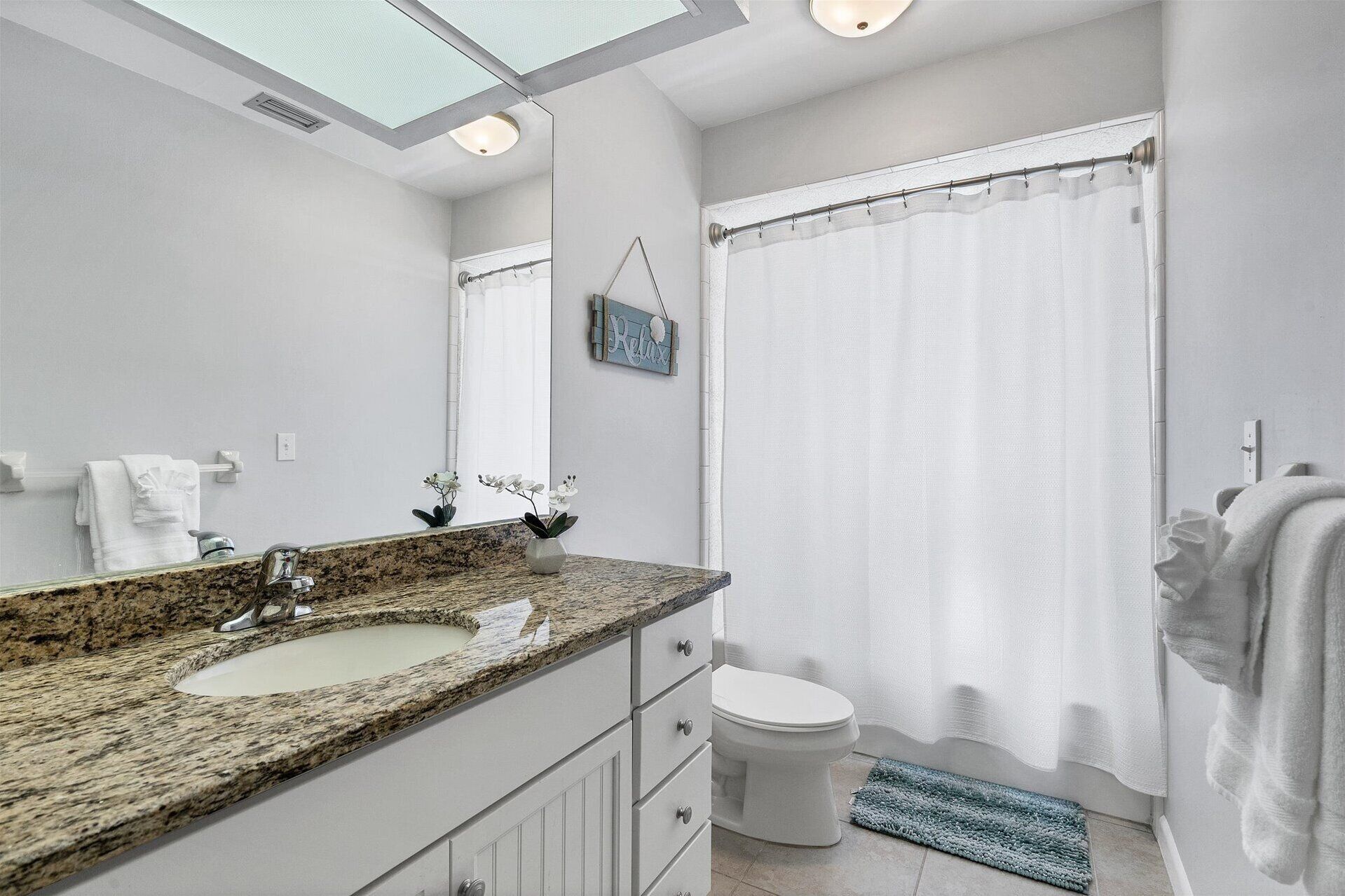 A bathroom with a granite countertop sink, a white toilet, a shower curtain, and a small rug.