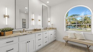 A modern bathroom with a large vanity, a large mirror, and a window with a view of palm trees and a pool.