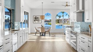 A modern kitchen with white cabinets, a dining area with a table and chairs, and a view of a pool and palm trees through large windows.