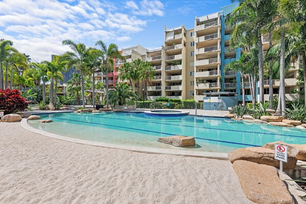 A resort pool with a sandy beach area, surrounded by palm trees and a multi-story building with balconies. - Resort-Style Comfort in the Heart of the Valley (Brisbane)