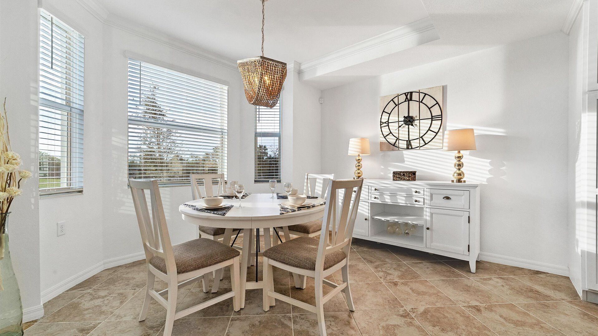 A dining area with a round table, chairs, a chandelier, a clock, and a cabinet with lamps.