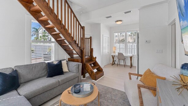 A spacious living room with a wooden staircase, a grey sofa, a coffee table, and a dining area with a chair and table. - Tay307-B (Punta Gorda)
