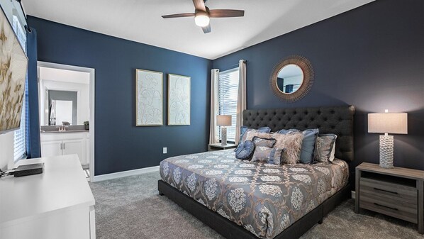 A bedroom with a tufted headboard, a large bed with patterned bedding, two framed wall art pieces, a round mirror, and a bedside table with a lamp.
