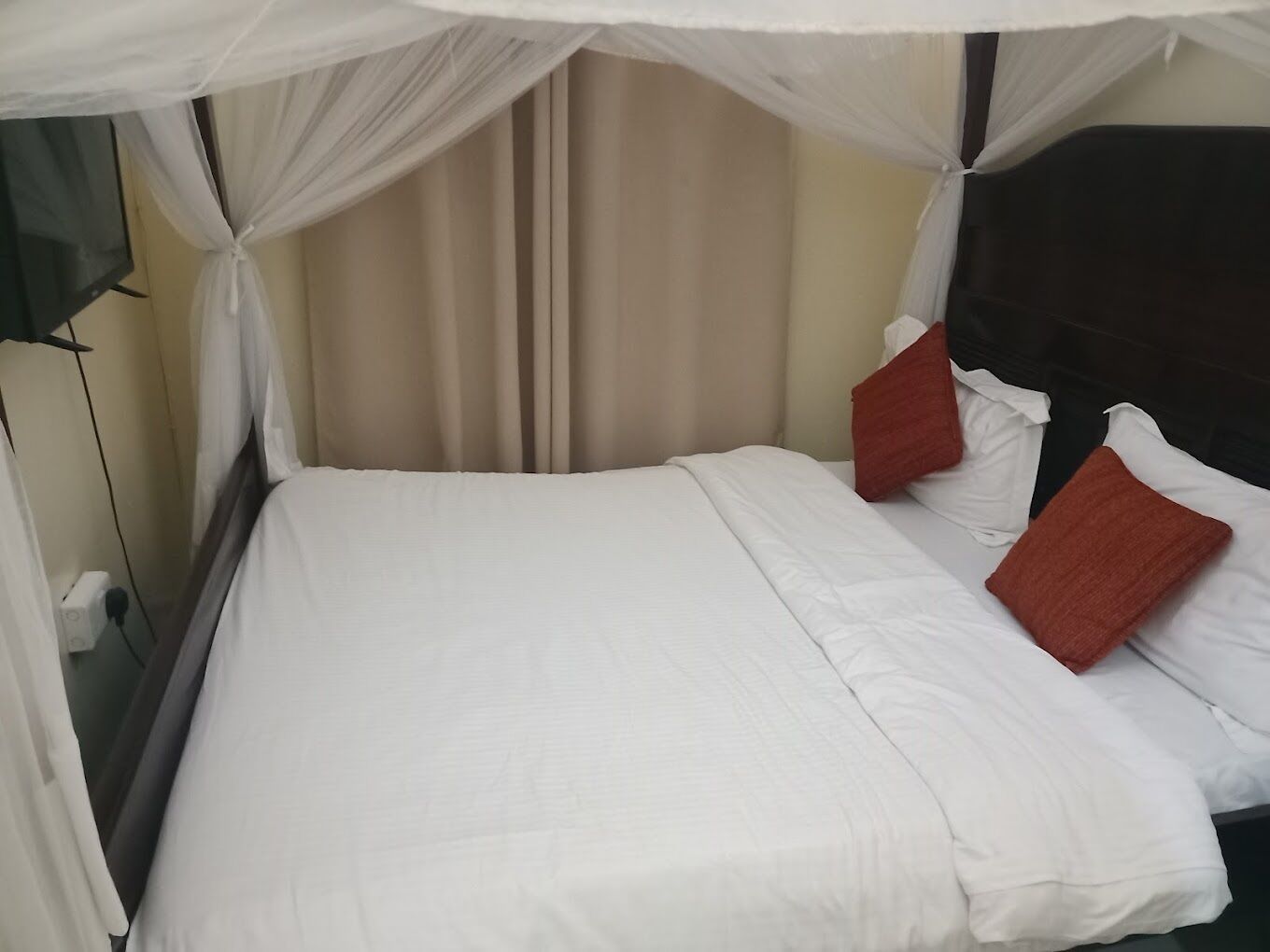 A neatly made bed with white bedding and two red pillows, a beige curtain, and a dark headboard.