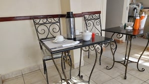Uma área de refeições com mesa e cadeiras de metal, uma cafeteira, xícaras e uma pequena mesa com temperos.