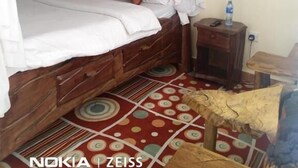 Uma cama com cabeceira de madeira, roupa de cama branca e um dossel. Há uma pequena mesa de cabeceira de madeira com uma garrafa de água e um controle remoto. O chão possui um tapete estampado.